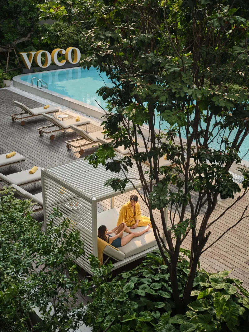 Couple relaxing in a poolside cabana at voco Surawong, with the VOCO sign and pool visible through the trees