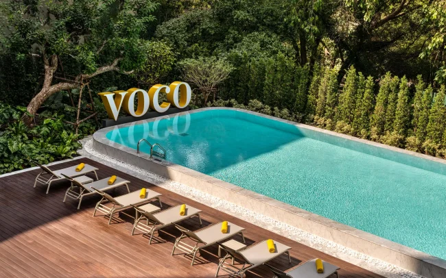 Empty pool deck with lounge chairs and the VOCO sign at voco Surawong, surrounded by tropical greenery