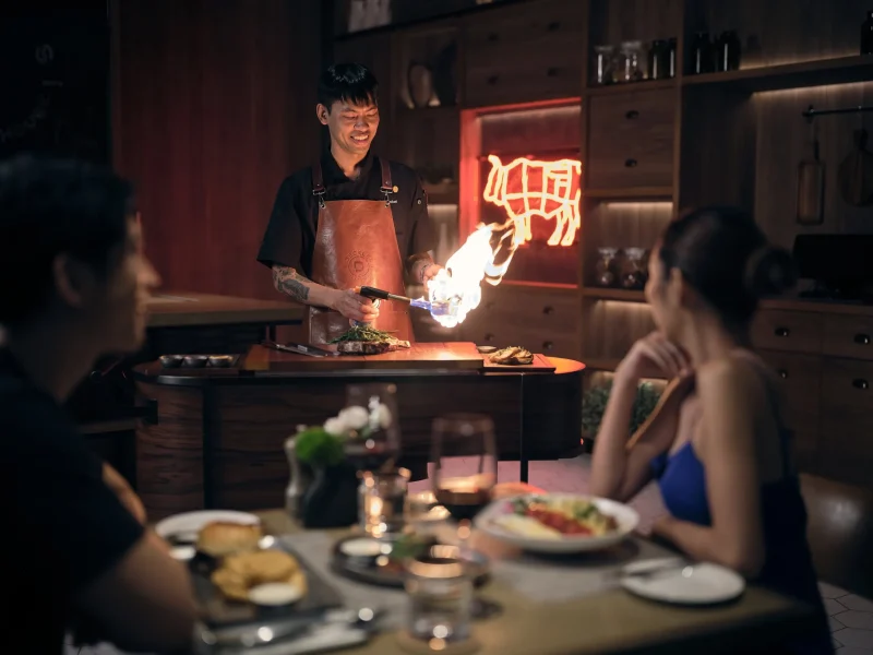 Chef torching a steak tableside at voco Surawong's steakhouse, with neon cow logo and diners watching