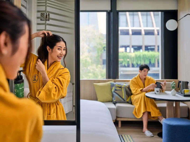 Woman in a yellow voco robe styling her hair in a Bangkok hotel room mirror, with man relaxing in the background